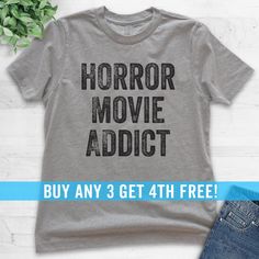 a gray shirt with the words horror movie addict on it, next to jeans and a plant