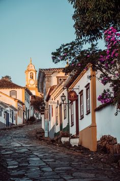 an old cobblestone street lined with white and yellow buildings in front of a clock tower