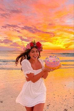 a woman in white dress holding a cake on beach with pink and yellow sunset behind her