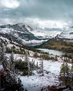 snow covered mountains and trees with water in the distance on a cloudy day near a lake