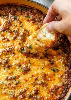 a hand holding a piece of bread over a casserole dish filled with cheese and meat