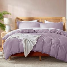 a bed with purple comforter and pillows in a pink room next to a potted plant