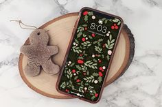 a teddy bear sitting next to an iphone case on top of a wooden slice with holly leaves and berries