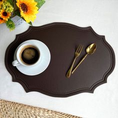 a cup of coffee sits on a placemat with gold spoons and fork next to it