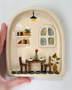 a hand holding up a ceramic plate with a miniature house on the front and shelves above it