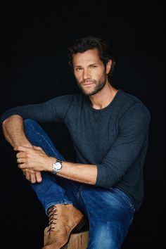 a man sitting on top of a wooden stool wearing jeans and a black t - shirt