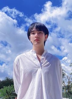 a young man standing in front of a blue sky with white clouds and trees behind him