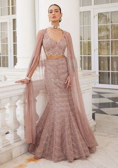 Editor's Note Elevate your style with our stunning salmon pink cape adorned with sequin and cutdana embellishments. The cape adds a touch of glamour and elegance to the ensemble. Paired with a scallop pattern fish cut lehenga and a sleeveless blouse, this outfit exudes sophistication and grace. The intricate detailing on the lehenga and blouse enhances the overall look, making it perfect for special occasions and weddings. Make a statement with this exquisite salmon pink ensemble and let your style shine. Fabric: Net Color: Pink Components: Lehenga, cape and blouse Occasion: Reception and engagement Note: Product colour may slightly vary due to photographic lighting sources Care: Dry clean only About the Designer Charu & Vasundhara's outfits epitomize the perfect blend of Indian and contem Jacket Blouse Lehenga, Jacket Lehenga Designer, Fancy Gowns Indian, Reception Lehenga Bridal Indian, Charu Vasundhara, Sangeet Outfit Bridal Gown, Reception Outfit For Bride Indian, Pink Sequin Lehenga, Reception Gown For Bride