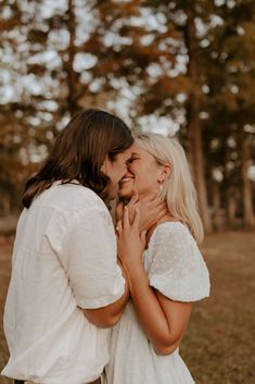 two women standing next to each other in front of trees and grass, one is kissing the other's cheek