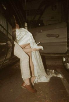 a woman in white dress sitting on the ground next to a car with her legs up