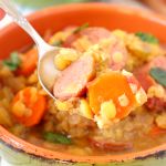 a spoon full of soup with carrots, corn and sausage on it in an orange bowl