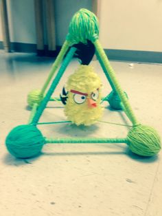 a yellow toy with green yarn on it's legs and eyes sitting in front of a ball of yarn