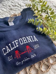 Cali, stand up! Comfy sweatshirt with California in a varsity-like font featuring the Golden Gate Bridge. This pullover Crewneck will keep you stylish and cool! You can add names on the sleeve for $5 extra!  This sweatshirt is available in 6 colors:  ✨ Forest Green ✨ Sand ✨ White ✨ Light Blue ✨ Navy  ✨ Heather Gray If there is a color you have in mind other than the ones available, send me a message and I can help! -Sweatshirt brand: Gildan -Sizes available: Adult Small - Adult 3XL -Fit: slightl Crew Neck T-shirt With Letter Embroidery For College, Embroidered Crew Neck Sweatshirt For School, Graphic Embroidered Crew Neck Sweatshirt For School, Crew Neck Sweatshirt With Embroidered Graphics For School, College Crew T-shirt With Embroidered Text, School Crew Neck Sweatshirt With Embroidered Text, School Sweatshirt With Embroidered Text And Crew Neck, Crew Neck Top With Embroidered Text For College, Collegiate Crew Sweatshirt With Embroidered Text