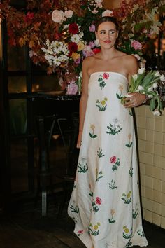 a woman in a white dress standing next to a wall with flowers and greenery