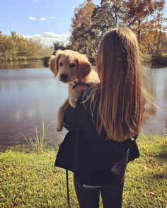 a woman holding a puppy in her arms near a lake with trees and grass around it