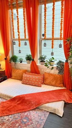 a bed with orange drapes and pillows in front of a window filled with potted plants