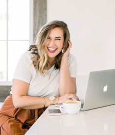 a woman sitting at a table with a laptop and coffee in front of her smiling