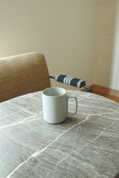 a white coffee cup sitting on top of a table next to a chair and window