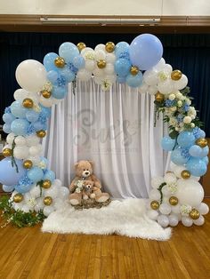 a teddy bear sitting in front of a white backdrop with blue and gold balloons on it