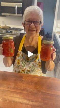 an older woman holding two jars of jam on top of a wooden table in a kitchen