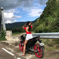 a woman sitting on top of a motor bike next to a road with mountains in the background