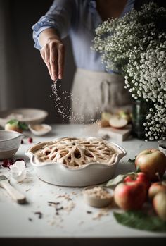 a person sprinkling sugar on top of a pie in a white dish next to apples