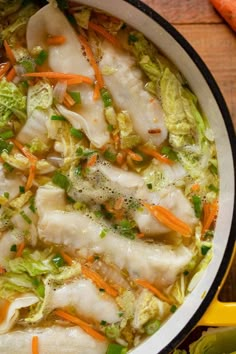 a pot filled with dumplings and vegetables on top of a wooden table next to carrots