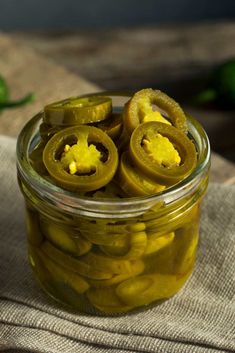 pickles in a glass jar sitting on a cloth next to green peppers and limes