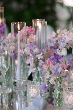 a table topped with lots of glass vases filled with purple and white flowers next to candles