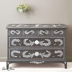 a black and white painted chest of drawers next to a plant hanging on the wall