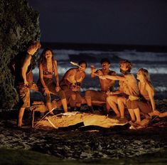 a group of people sitting around a campfire on the beach at night with torches