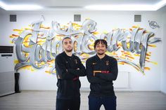 two men standing next to each other in front of a wall with graffiti on it