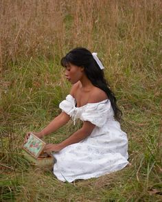 a woman in a white dress sitting on the ground reading a book with her hands