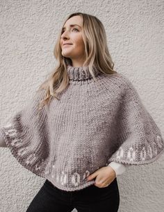 a woman standing in front of a white wall wearing a gray sweater and black pants