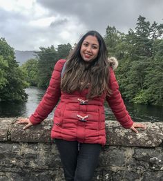 a woman standing next to a stone wall with trees in the background on a cloudy day