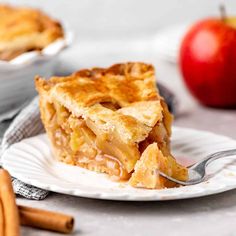 a slice of apple pie on a white plate with cinnamon sticks and apples in the background