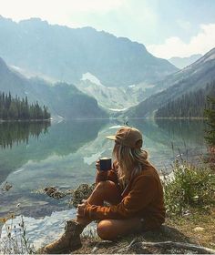 a woman sitting on the edge of a lake holding a coffee cup in her hands