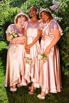 three women in pink dresses standing next to each other on the grass with flowers and bushes behind them