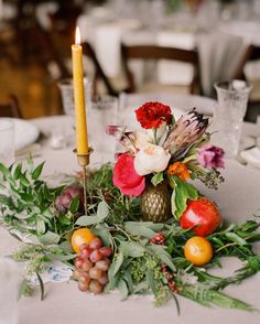 an arrangement of fruit and flowers on a table with a candle in the centerpiece