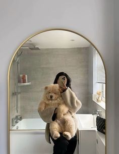 a woman holding a teddy bear in front of a bathtub with a mirror on the wall