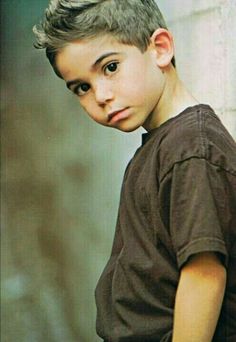 a young boy standing next to a wall with his hand on his hip and looking at the camera