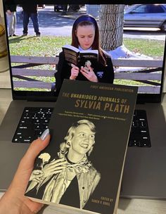 a person is reading a book in front of a laptop and computer screen with a woman's face on it