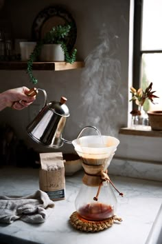 a person pours coffee into a glass teapot on a kitchen counter with other items