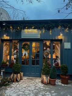 a store front with potted plants and wreaths on the windows at night time