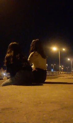 two people sitting on the ground at night with their backs to each other and one person looking off into the distance