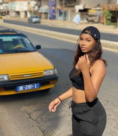 a woman standing in front of a yellow car on the side of the road with her hand under her chin