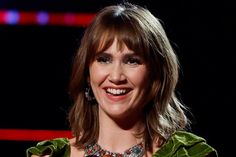 a smiling woman in a green jacket and necklace on stage with red lights behind her