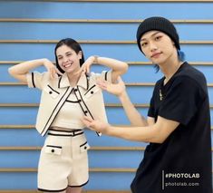 two people standing next to each other in front of a blue wall and one person making the peace sign with their hands