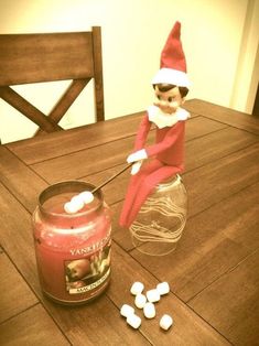 an elf sitting on top of a wooden table next to a jar filled with marshmallows