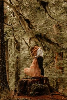 a man and woman standing on top of a tree stump in the forest hugging each other